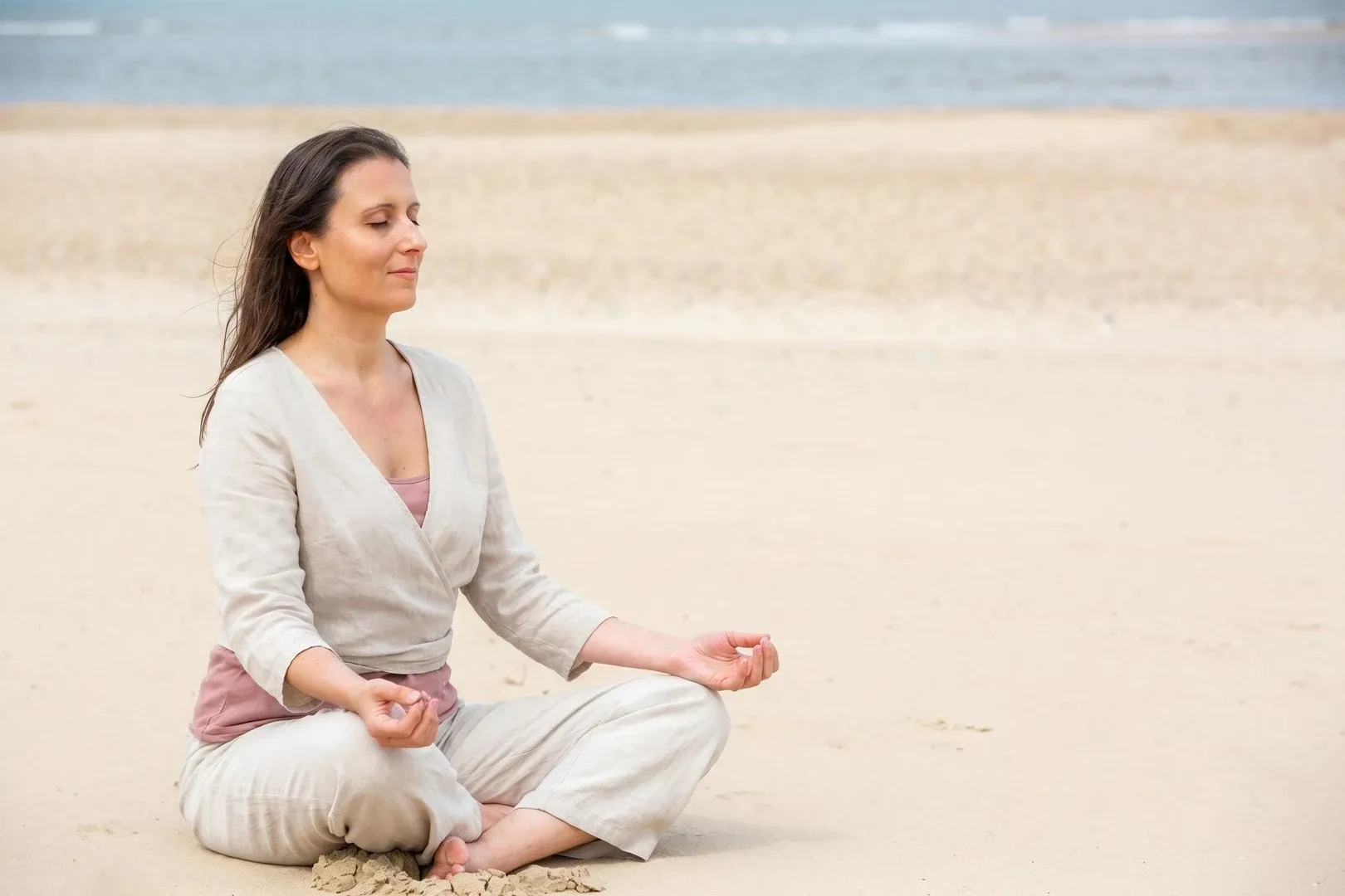 Ania seated cross-legged in meditation on warm pale sand by a calm sea