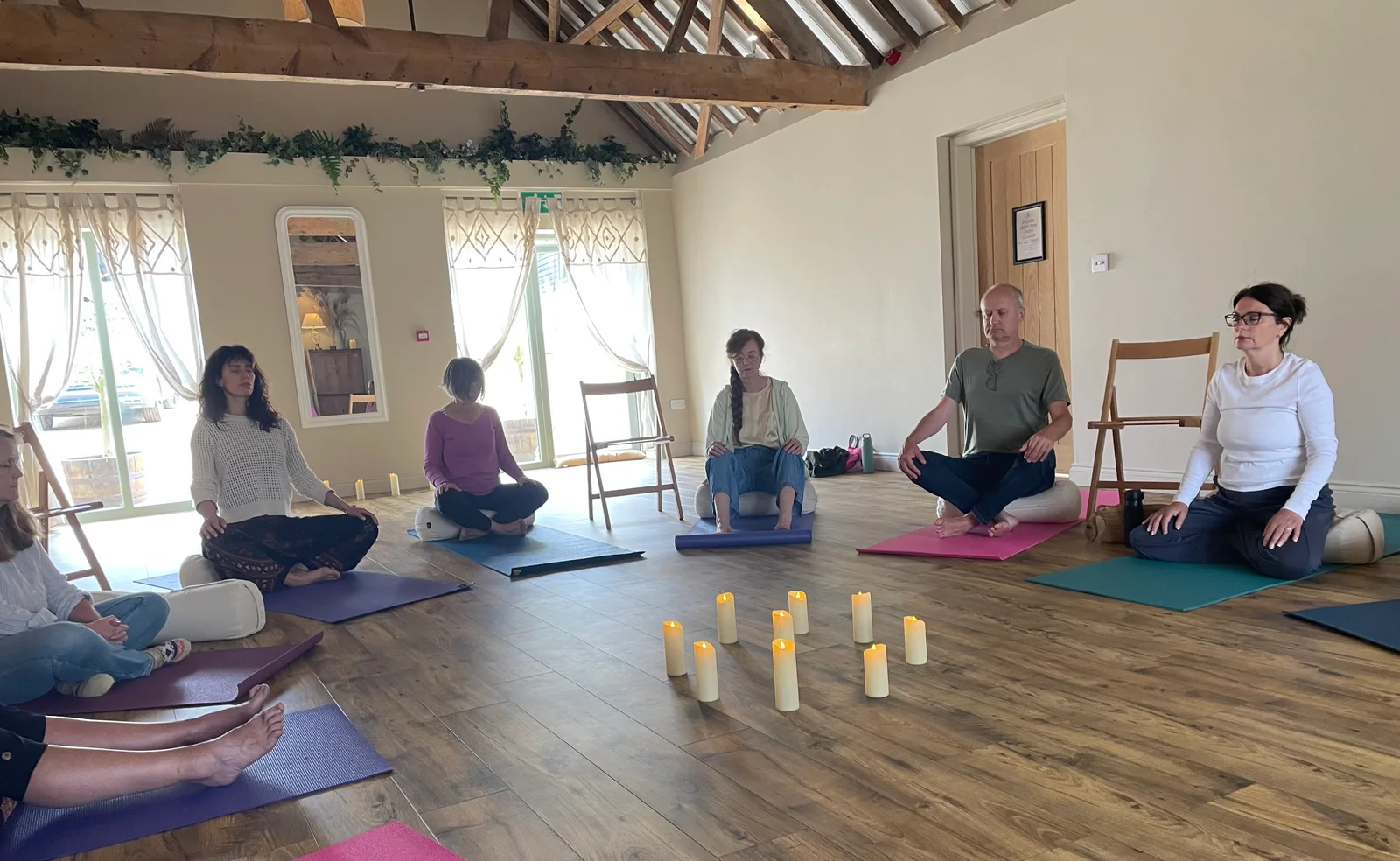 Participants sitting in meditation on yoga mats in a circle around a cluster of candles, in the light-filled room at The Painted Barn